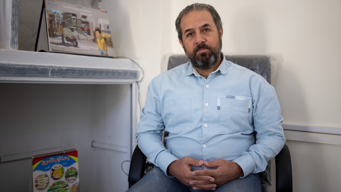 Un homme est assis sur une chaise. Il porte une chemise bleu clair à manches longues et un pantalon sombre. À gauche de l’image, une étagère murale contient plusieurs objets.