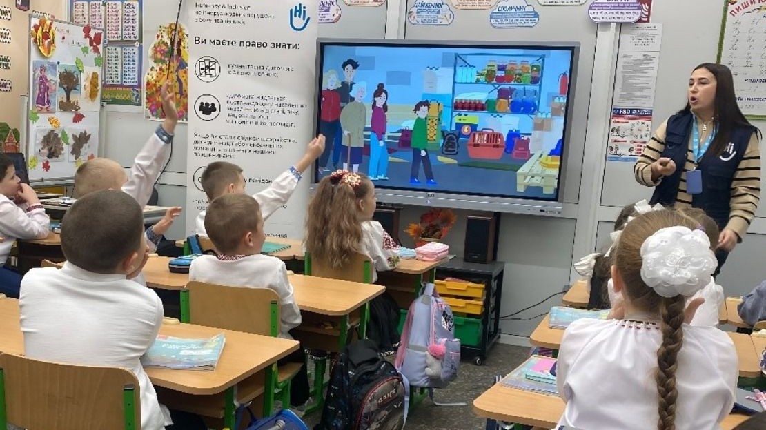 Des enfants sont assis à leurs bureaux dans une classe face à un écran où l'on montre des images pédagogiques, une famille ukrainienne dans sa vie de tous les jours pour expliquer les comportements sûrs à adopter en cas de danger. Au tableau, l'agent de sensibilisation de HI se tient debout et leur décrit les images.
