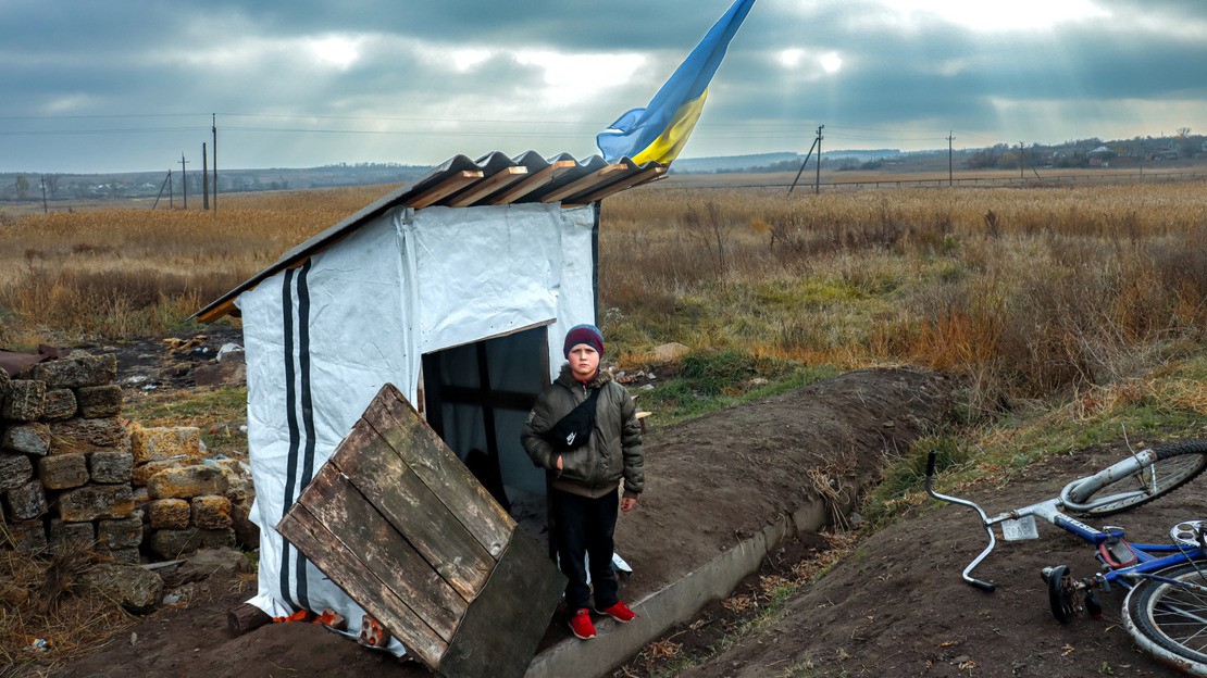Un jeune garçon d'une dizaine d'années se tient debout devant une cabane arborant le drapeau ukrainien, au milieu d'un champ. Sur la droite, son vélo est couché sur le côté.