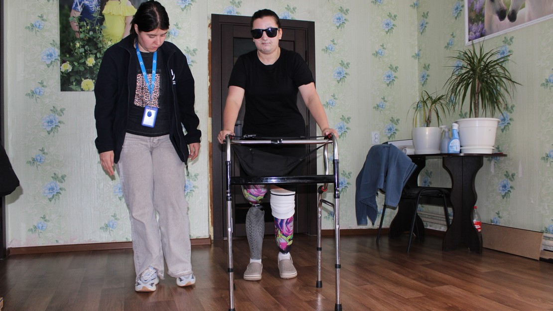 Aidée d'une travailleuse humanitaire, une femme qui porte des lunettes noires marche avec un déambulateur. Elle est amputée des deux jambes et est équipée de prothèses.