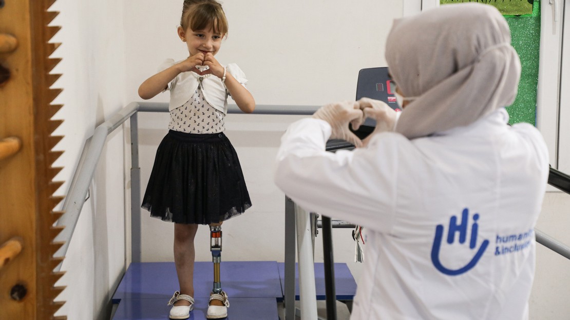 Une fillette souriante équipée d'une prothèse de jambe gauche forme un cœur avec ses mains en direction d'une femme, de dos au premier plan, qui lui renvoie son geste.